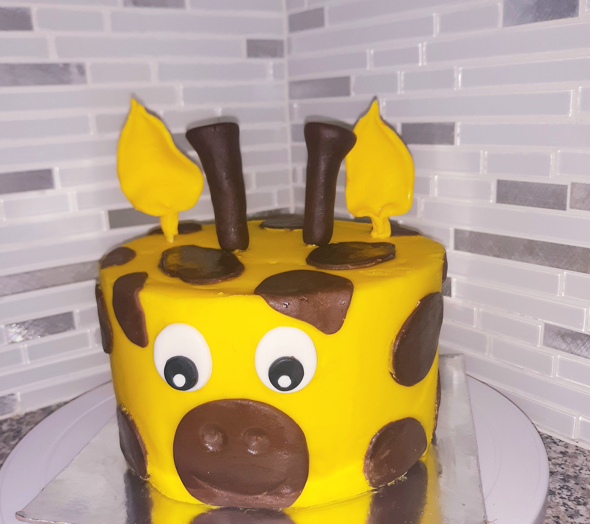 Yellow cake with brown decorations resembling a cow on a white plate against a tiled wall background.